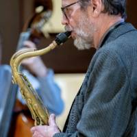 Man plays the saxophone with man playing cello in background